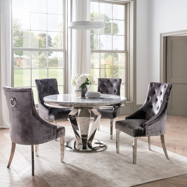 lavish_ A modern dining area in Southport with an Arturo Round Marble Dining Table, four tufted velvet chairs, and a view of the garden through large windows.