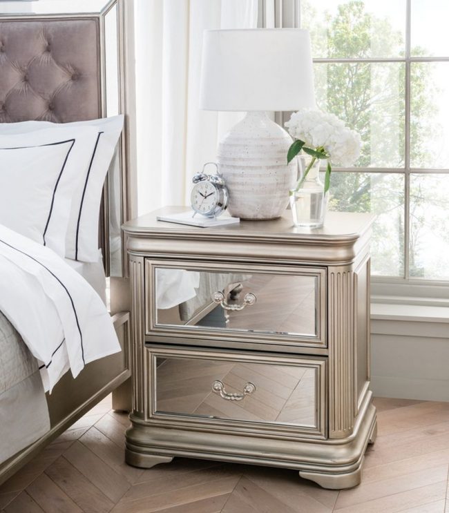 lavish_ Elegant Jessica bedside table with a lamp, clock, and vase of flowers next to a bed with striped linens.