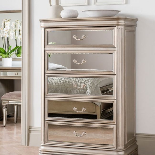 lavish_ Sentence with product name: An elegant Jessica 5 Drawer Tall Chest with partially open drawers, featuring ornate handles and decorative detailing, stands as a statement piece of furniture in a Southport room with light-toned decor.