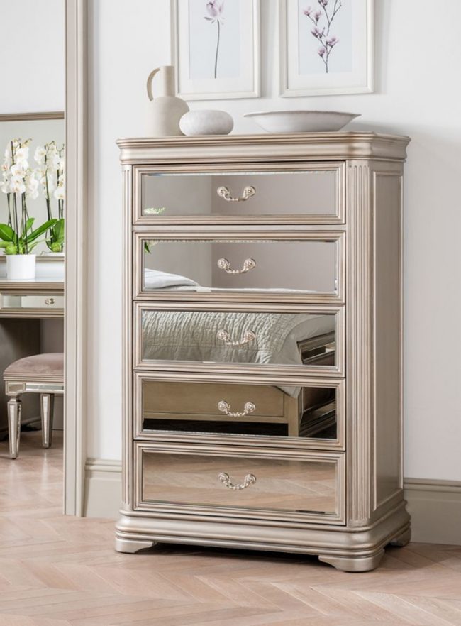 lavish_ Sentence with product name: An elegant Jessica 5 Drawer Tall Chest with partially open drawers, featuring ornate handles and decorative detailing, stands as a statement piece of furniture in a Southport room with light-toned decor.